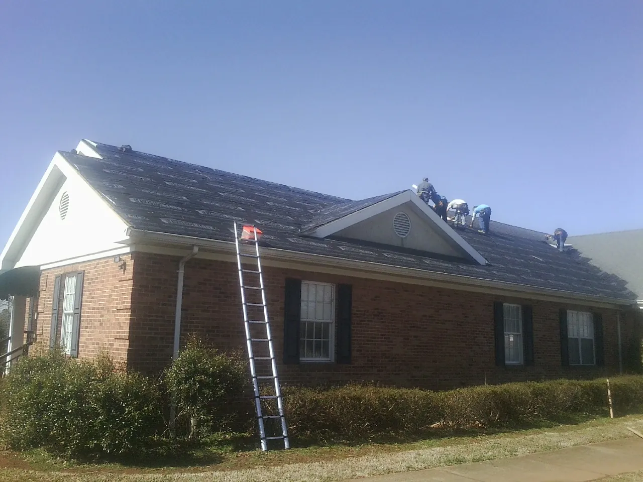 Crew tearing shingles off a brick ranch roof with a ladder set — Metal Roof Repair work by our crew in Howell